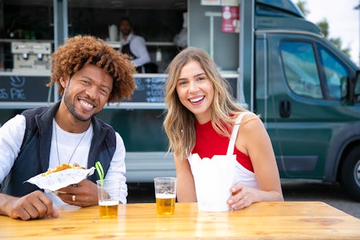 Cheerful young lady with African American male friend smiling and looking at camera while sitting at table in street cafe and eating burger with french fries and drinking beer